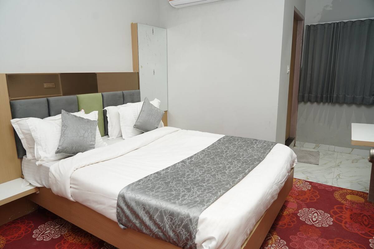 Una cama bien hecha con una colcha gris y blanca estampada, un cabecero de madera y una alfombra roja con diseño.