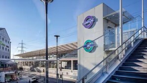 Una estación de tren moderna con cielo despejado, una zona de espera techada y una escalera que conduce a un nivel superior.