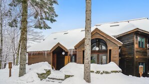 A wooden cabin with a snow-covered roof, surrounded by trees and a snow-covered ground. - One-bedroom condo with pool, hot tub, weekend winter, shuttle, and EV charger (Killington)