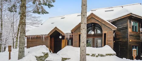 A wooden cabin with a snow-covered roof, surrounded by trees and a snow-covered ground.