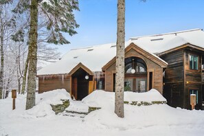 Une cabane en bois, dont le toit est recouvert de neige, entourée d’arbres et posée sur un sol enneigé.