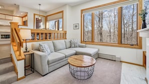 A modern living room with a grey sectional sofa, a round coffee table, and a large window offering a view of trees. - Cozy Highridge Condo Unit I6 in Killington with pool and hot tub. (Killington)