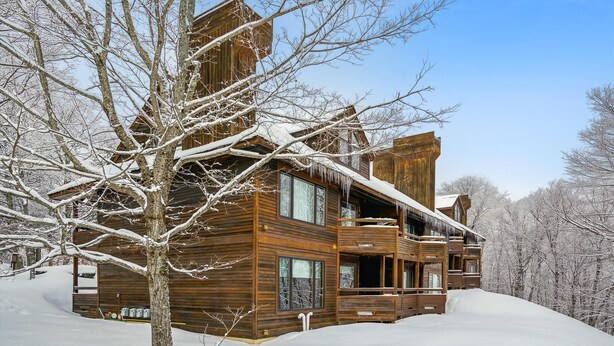 A wooden cabin with a large porch and multiple windows, surrounded by snow-covered trees. - Cozy Highridge Condo Unit I6 in Killington with pool and hot tub. (Killington)
