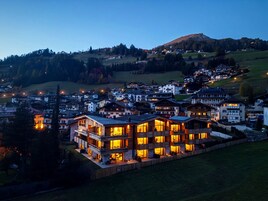 Un immeuble à plusieurs étages doté de balcons, entouré d’une zone verdoyante et avec un paysage vallonné en arrière-plan.