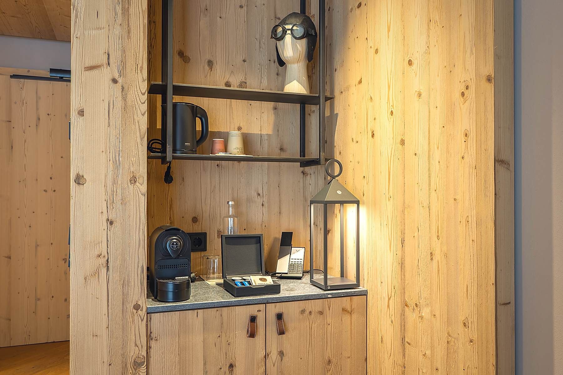 A wooden interior with a shelf holding a coffee maker, a lamp, and a small table with a laptop and a glass.