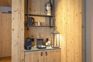 A wooden interior with a shelf holding a coffee maker, a lamp, and a small table with a laptop and a glass. - La Tambra - Hotel Lodge (Santa Cristina Val Gardena)