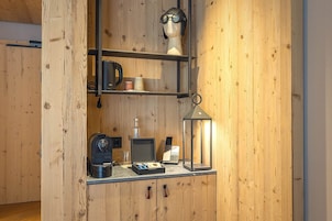 A wooden interior with a shelf holding a coffee maker, a lamp, and a small table with a laptop and a glass.