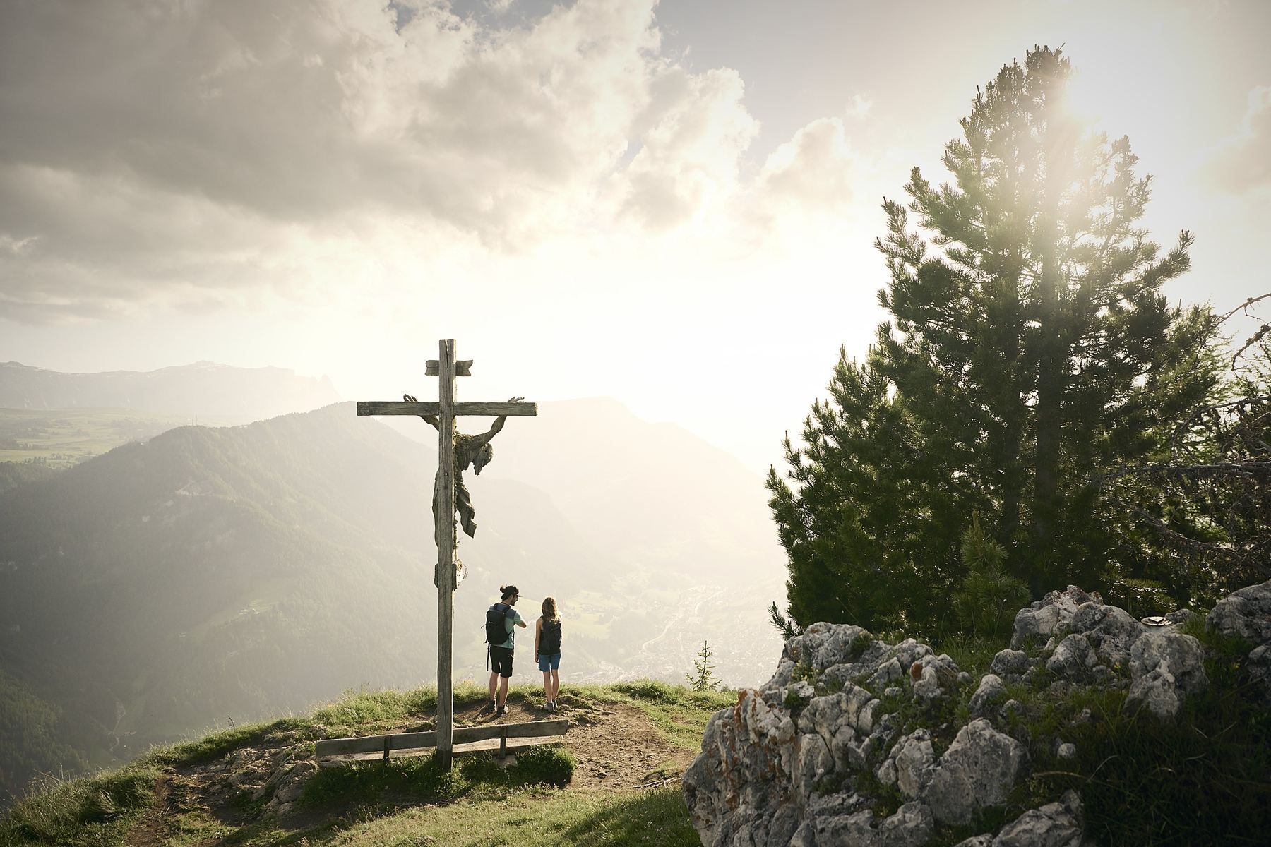 A wooden cross on a hill with two people standing nearby, surrounded by mountains and a pine tree.