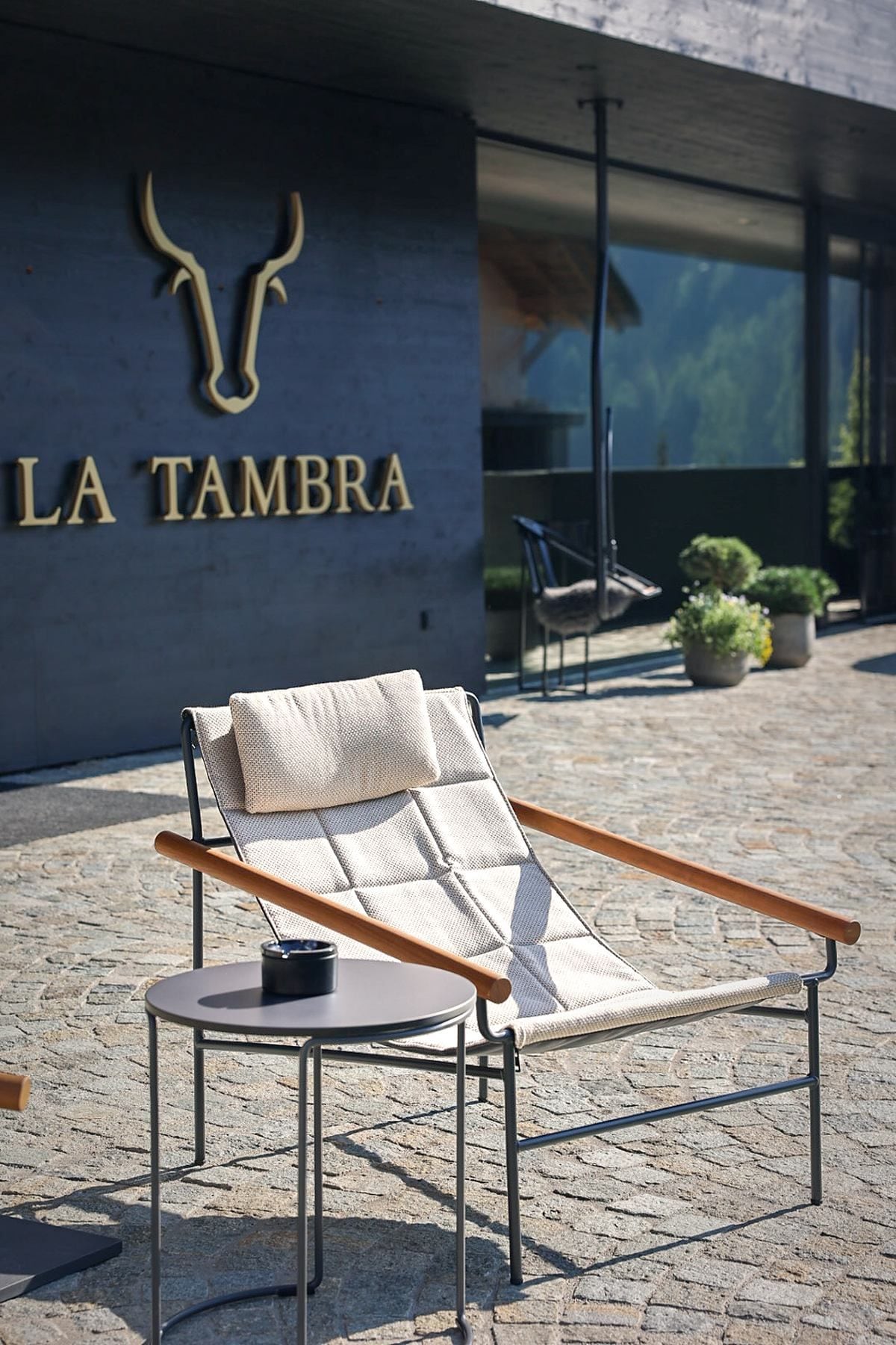 A lounge chair with a cushion and a small table, positioned in front of a building with a sign that reads 'LA TAMBRA'.