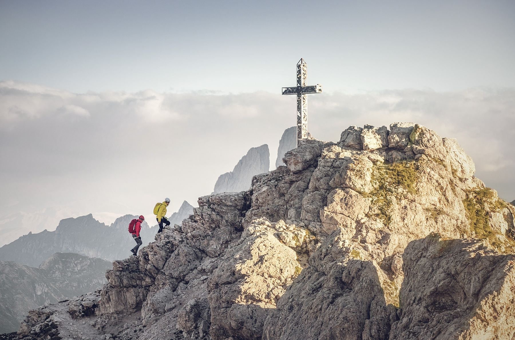 A rugged mountain peak with a cross structure on top and hikers at the base.