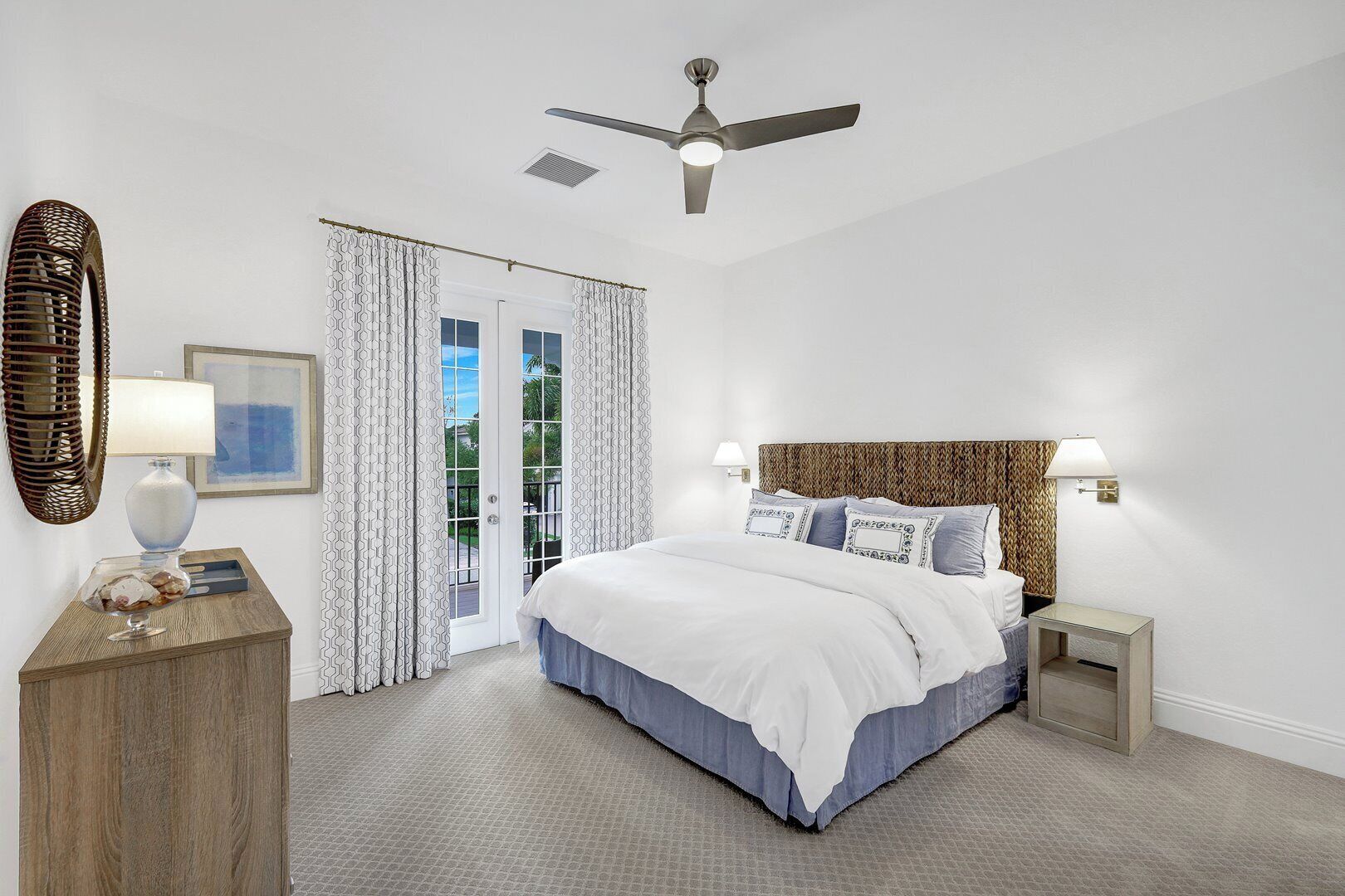 A bedroom with a bed, bedside lamps, a ceiling fan, a door with patterned curtains, and a small table with a lamp and decorative items.