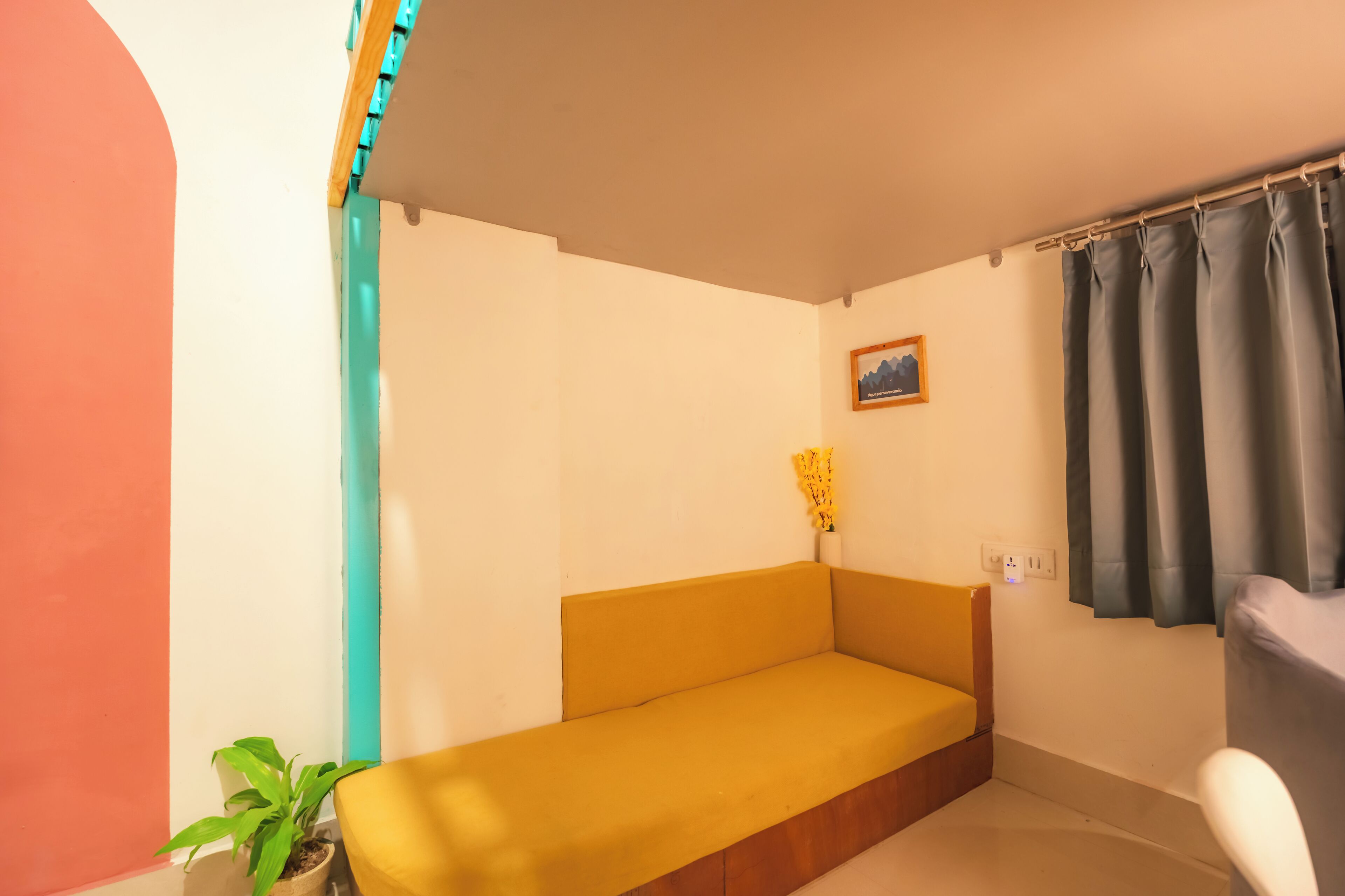 Ein Zimmer mit einem gelben Sofa, einem Topf mit Pflanze, einem gerahmten Bild an der Wand und einem Fenster mit Vorhängen.