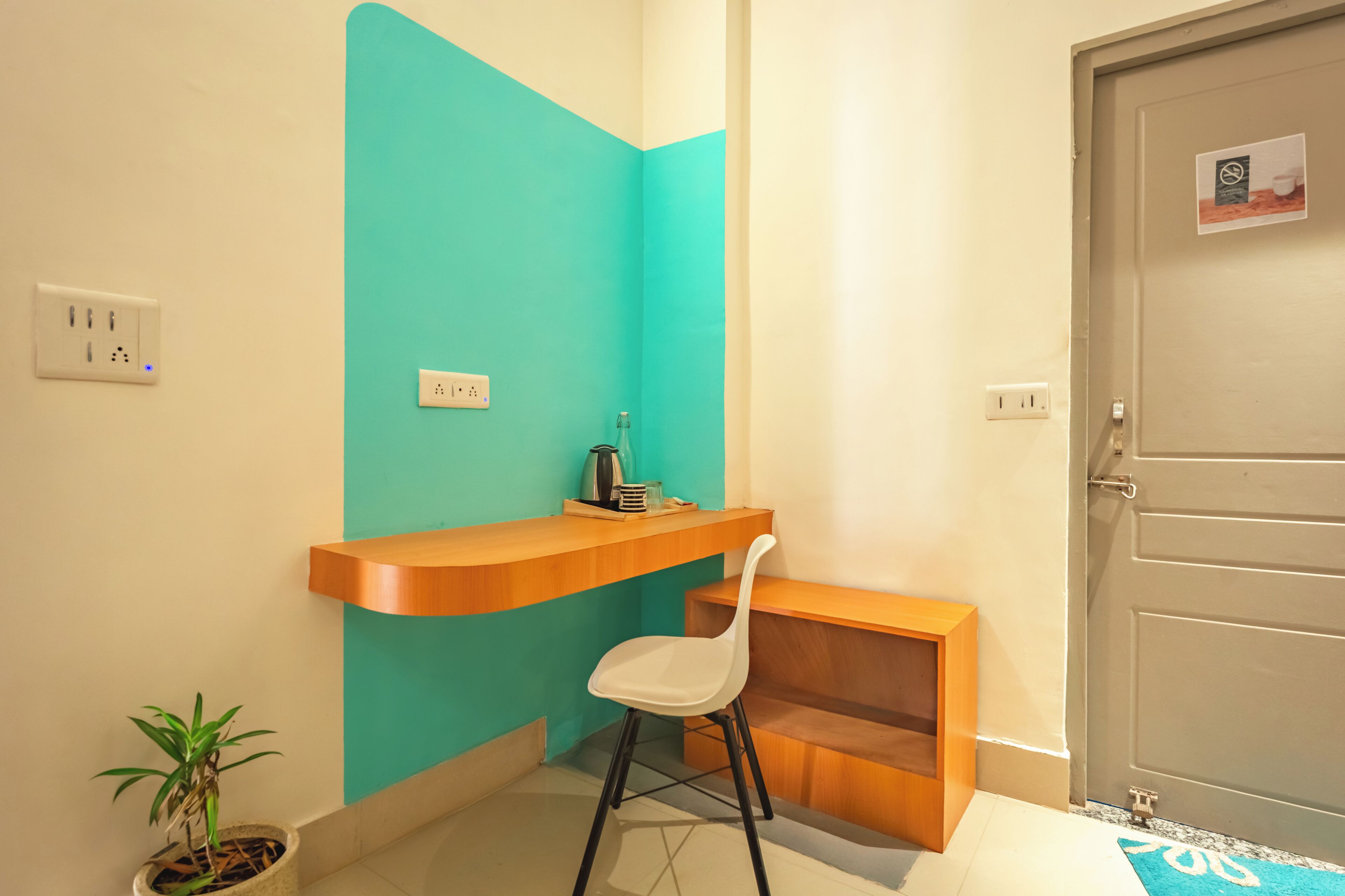 A small, modern room with a teal accent wall, a wooden desk, a white chair, a potted plant, and a door with a sign.