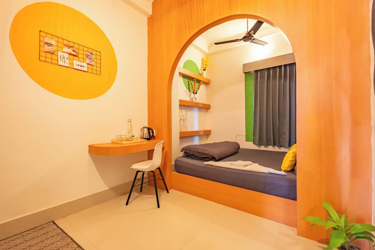 Une pièce de taille modeste comprenant un lit, un bureau et une chaise. On y trouve une petite étagère avec des objets décoratifs et une plante.