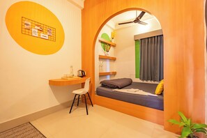A compact room with a bed, a desk, and a chair. There is a small shelf with decorative items and a plant.