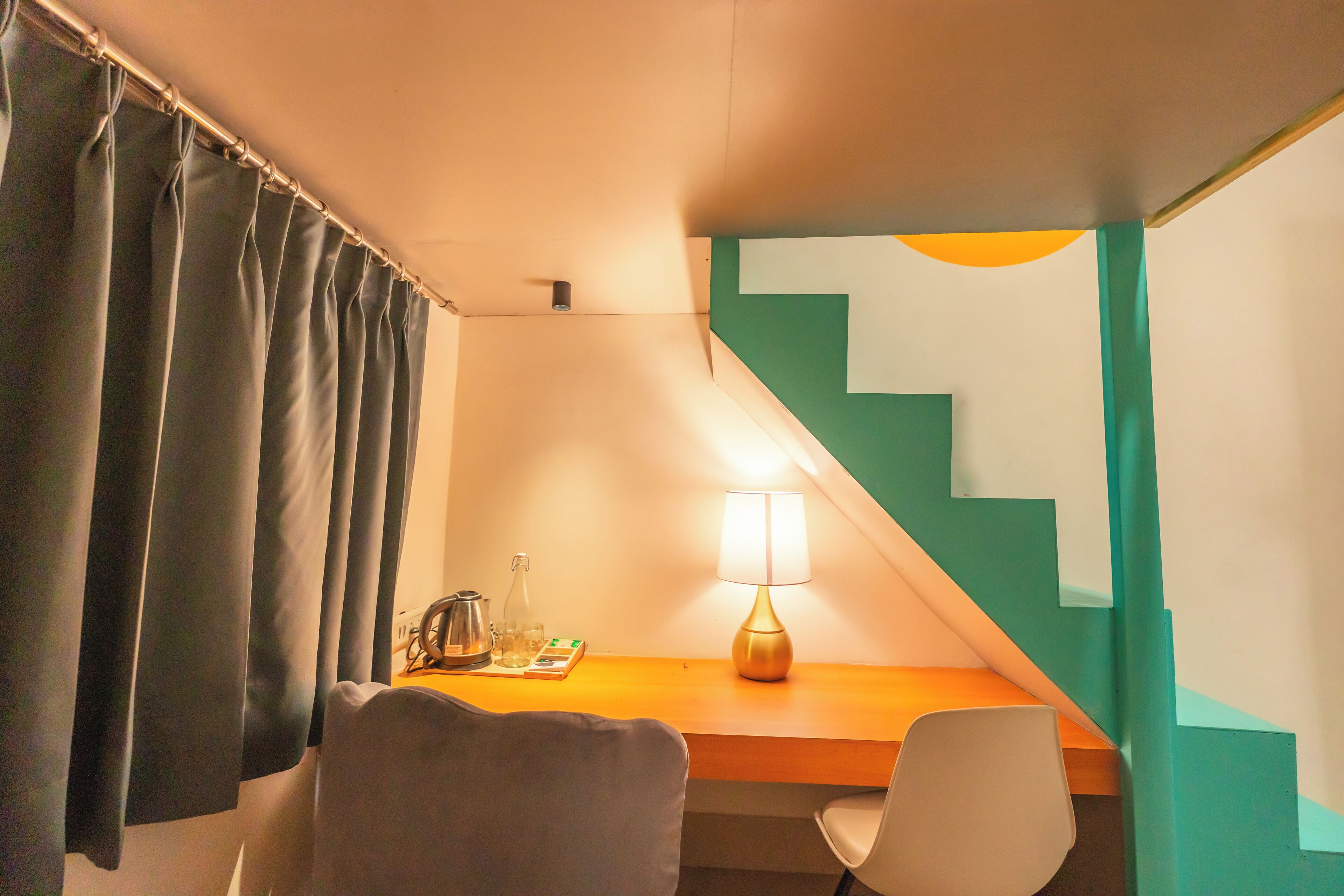 A hotel room with a desk, chairs, a lamp, a coffee maker, and a staircase with teal accents.