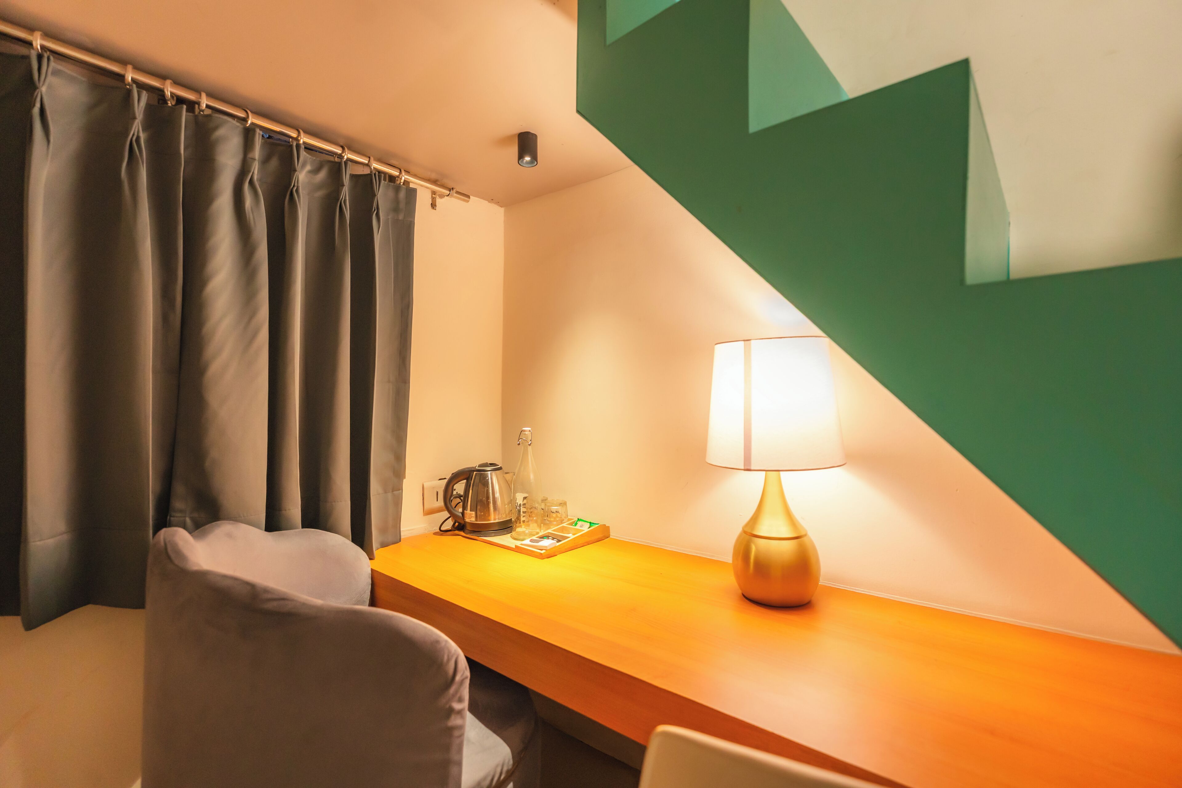 Ett hotellrum med ett skrivbord i trä, en lampa, en stol, en kaffebryggare och en gardin.