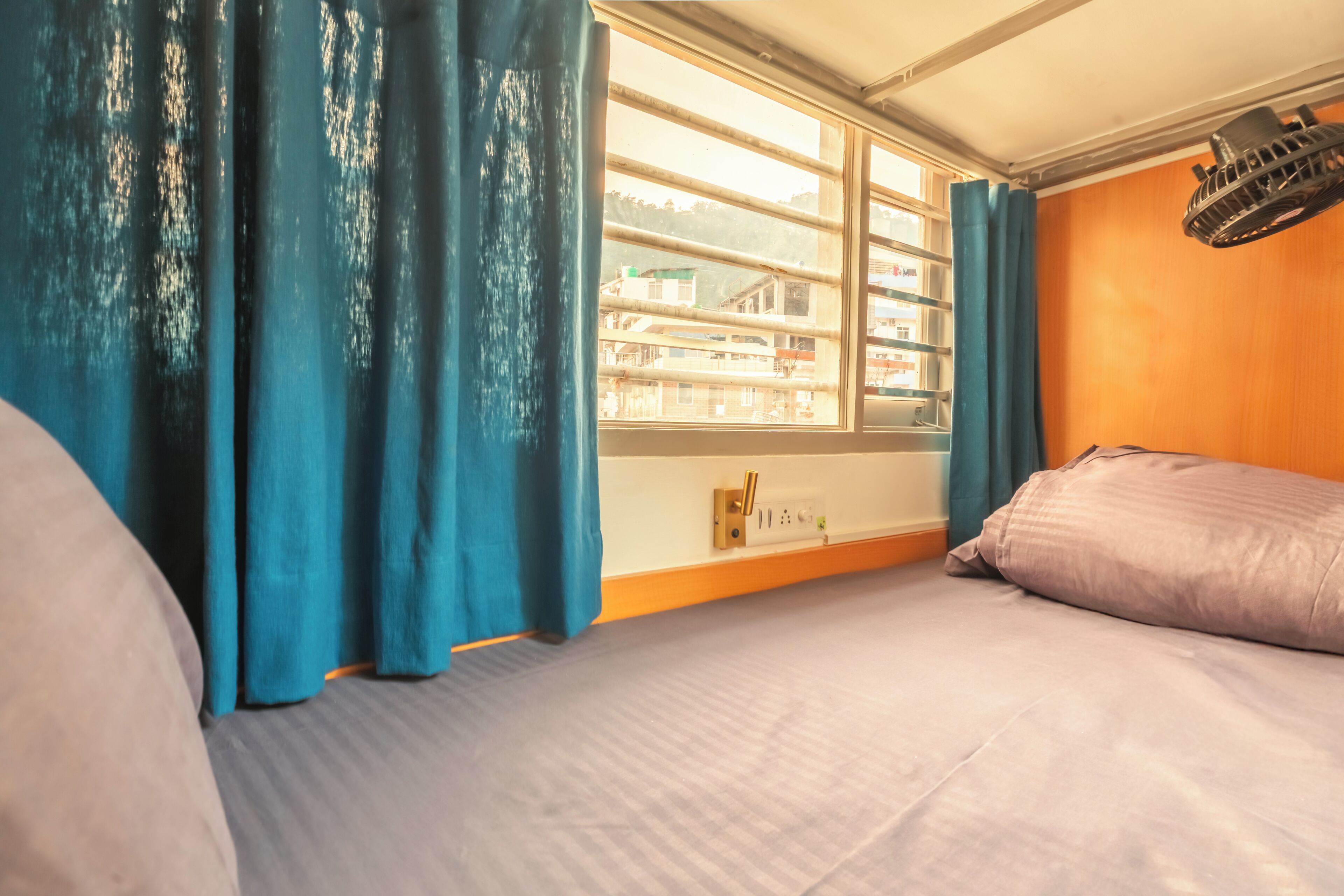 A room with a bed, a window with curtains, a wall-mounted heater, and a view of buildings outside.