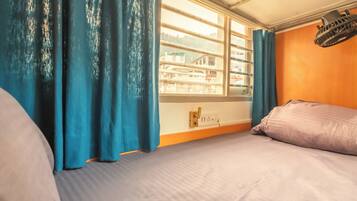 A room with a bed, a window with curtains, a wall-mounted heater, and a view of buildings outside.