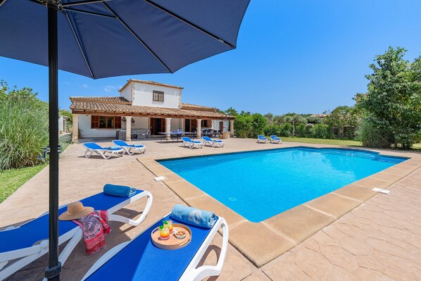 Una piscina con sillas de descanso y sombrillas, una casa con techo de tejas y un jardín.