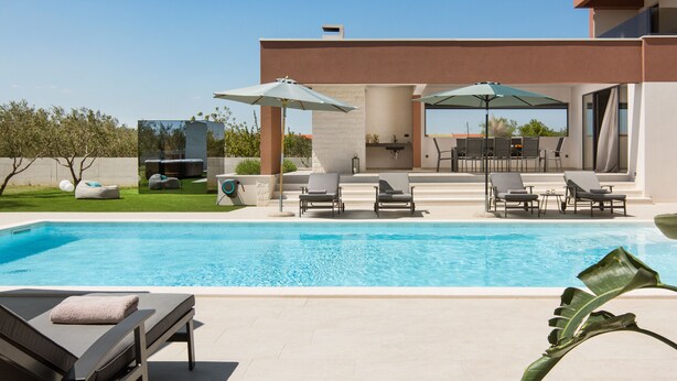 Un área de piscina moderna con sillones de playa y sombrillas, cielo despejado y un césped bien cuidado.