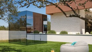 Un espacio exterior moderno con estructura de vidrio, un sillón informal de color gris y una pequeña mesa con una bebida.
