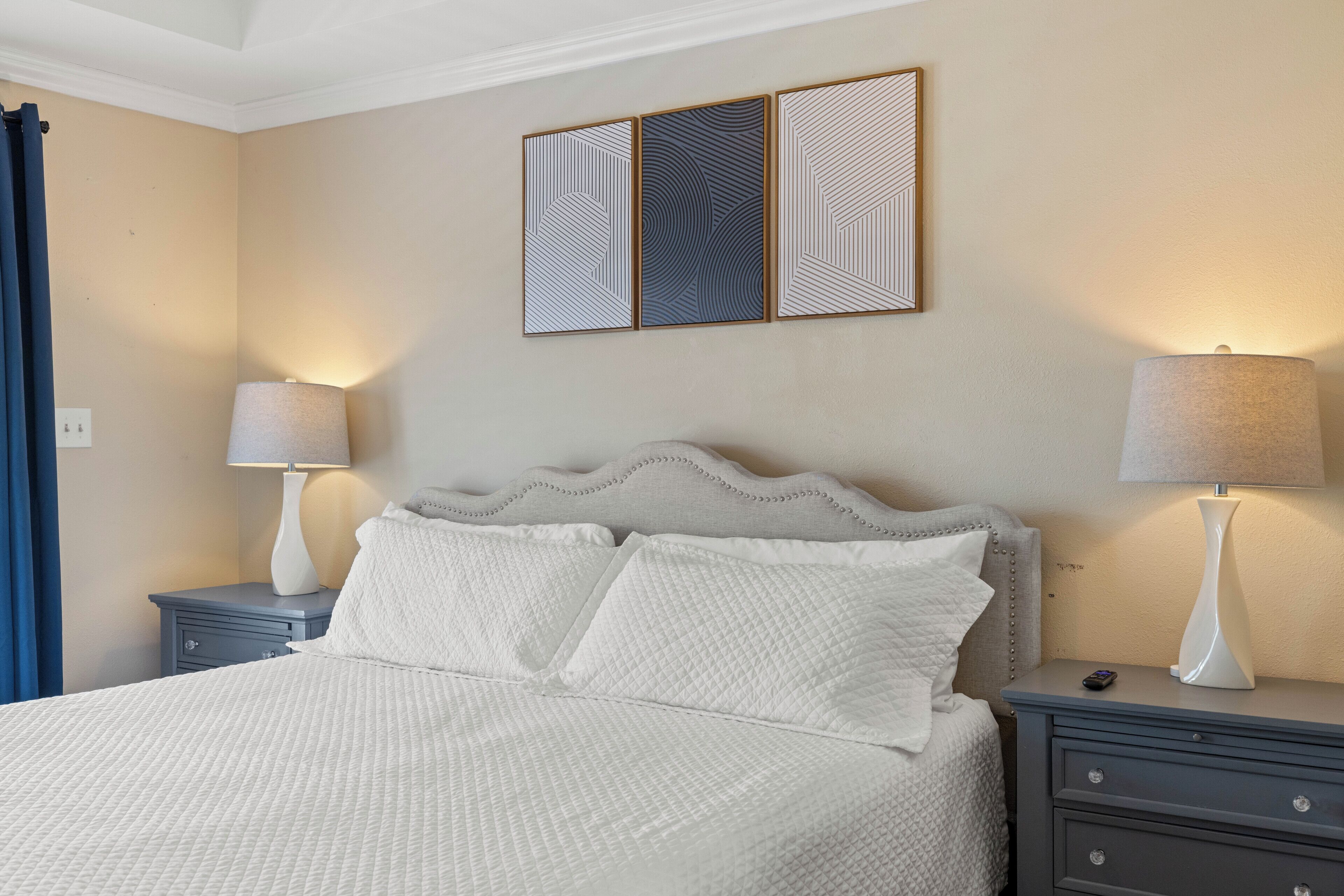 A neatly made bed with a textured white duvet, a headboard with a decorative pattern, two bedside tables with lamps, and a wall-mounted artwork above the bed.