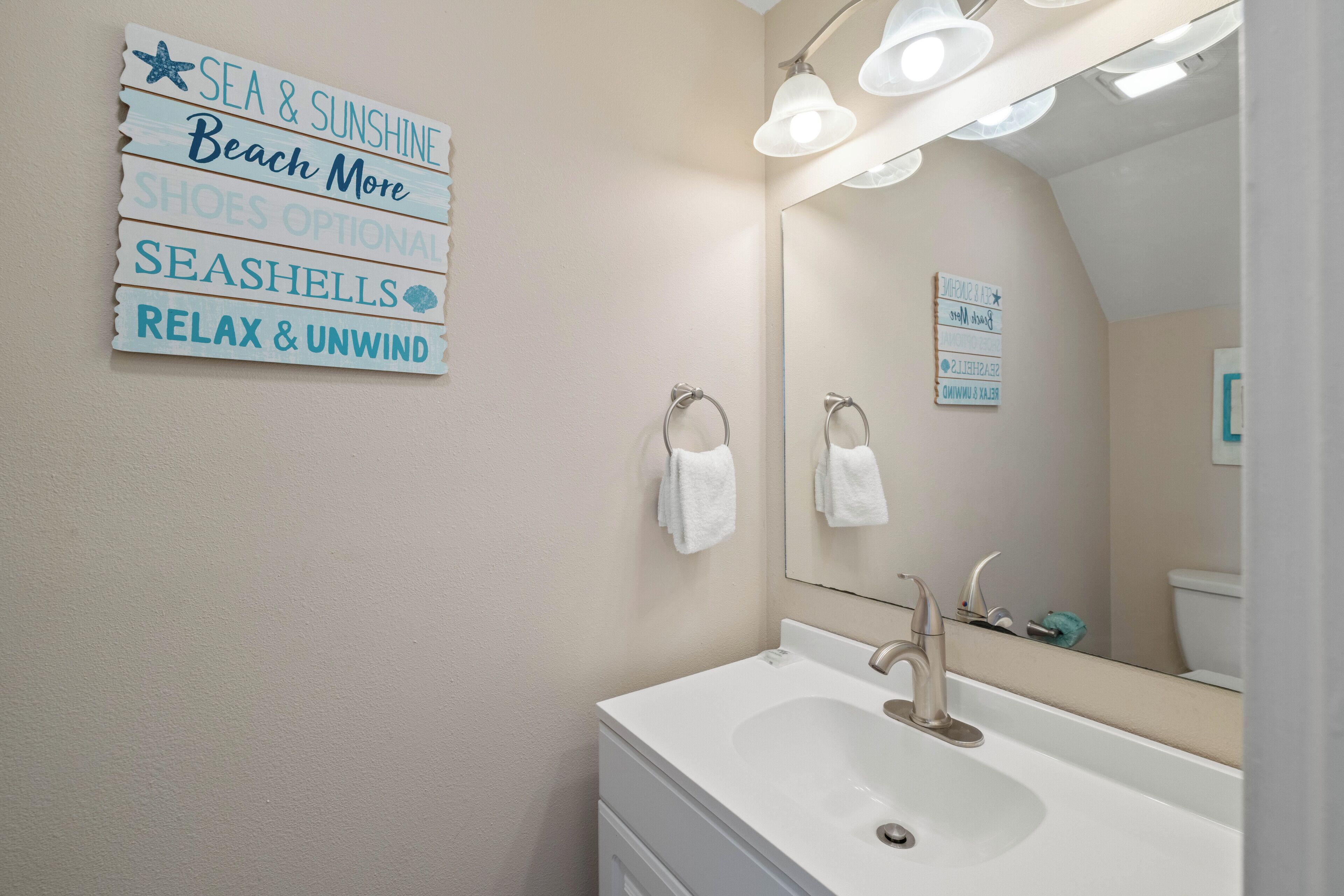 Une salle de bain équipée d’un lavabo, d’un miroir et d’un porte-serviettes. Un panneau avec du texte est accroché au mur.
