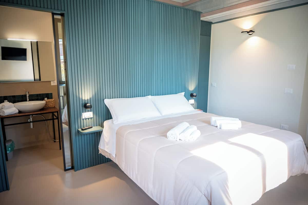 Habitación de hotel con cama, mesita de noche, espejo y baño con lavamanos y toallero.