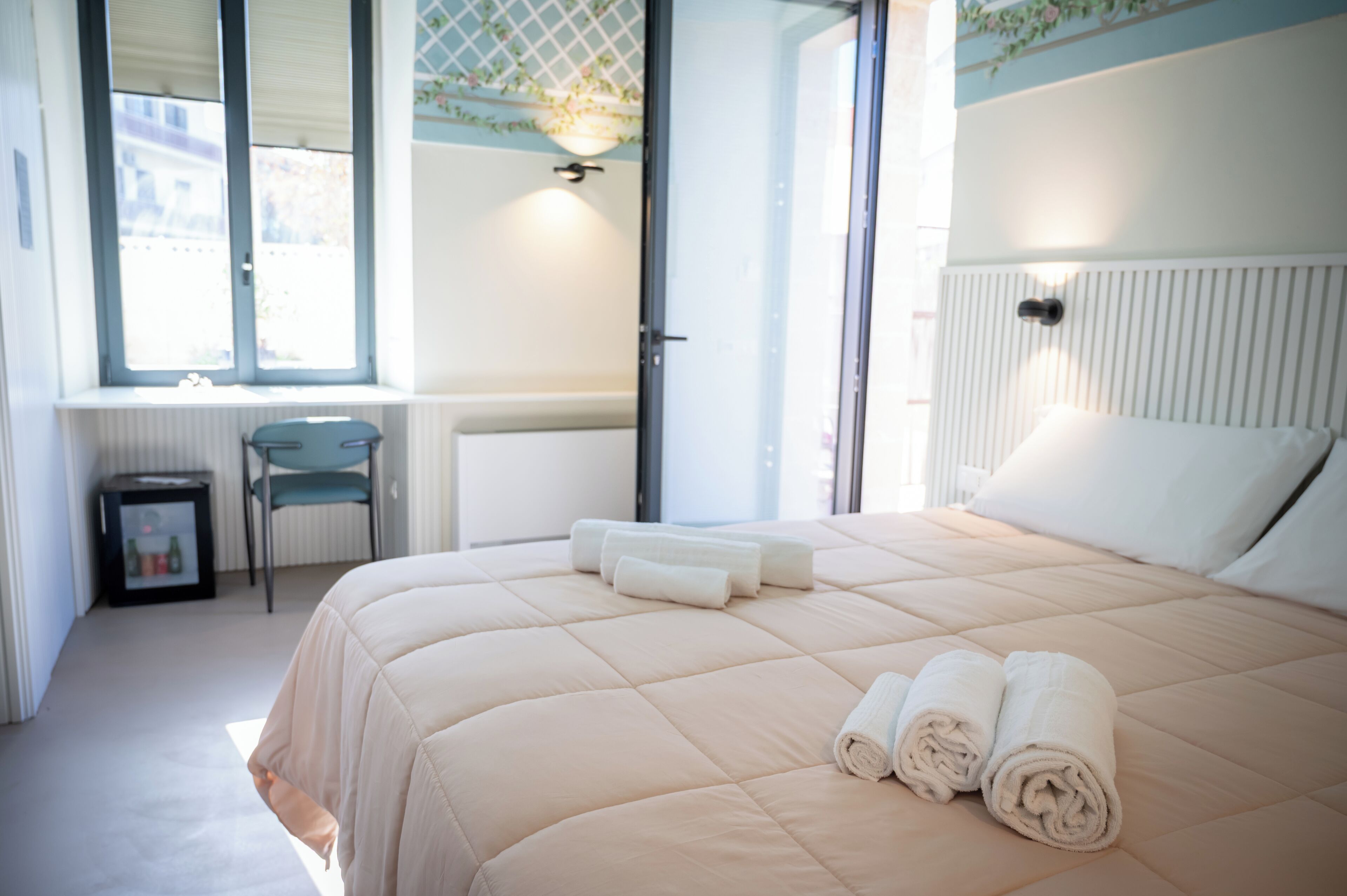 Una cama bien hecha con toallas enrolladas, una pequeña mesa con una silla y una ventana con persianas.