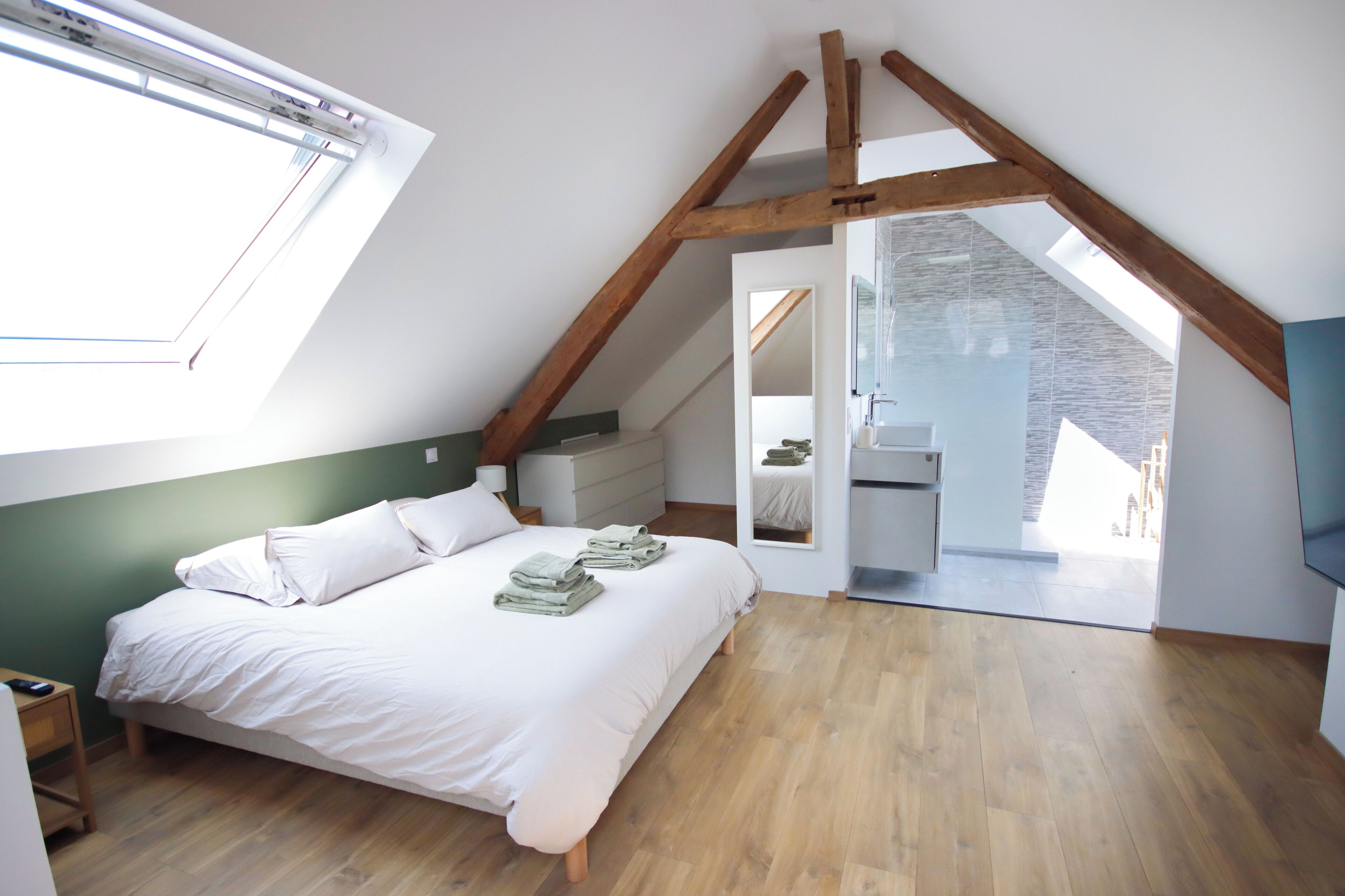 A bedroom with a sloped ceiling, a bed with white linens, a wooden floor, and a view of a bathroom through an open door.