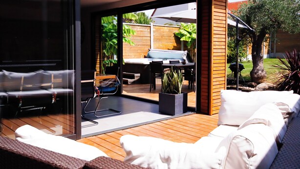 A modern outdoor patio with a wooden deck, white cushioned seating, and a glass door leading to a garden area with a barbecue and outdoor furniture.