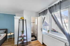 Een kamer met een bed, een klein tafeltje met een lamp, een plank met planten, een raam met gordijnen en uitzicht op een badkamer.