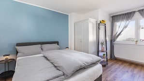Une chambre à coucher avec un lit, une table de chevet, une armoire, une fenêtre avec des rideaux et une plante posée sur une étagère.