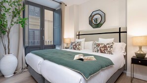 Ein Hotelzimmer mit einem großen Bett, einer Zimmerpflanze, einem Nachttisch und einem Fenster mit Vorhängen.