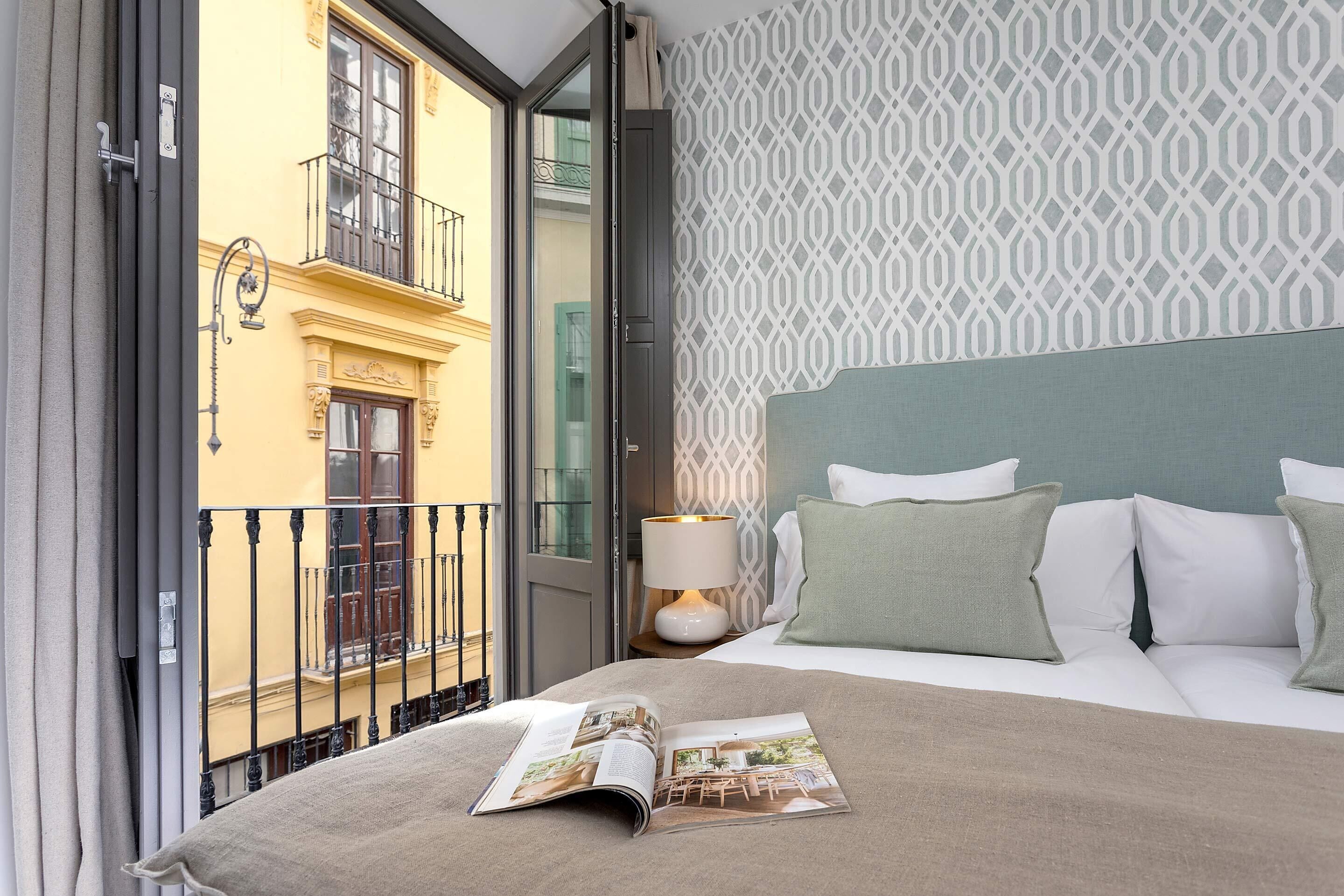 Une chambre d’hôtel avec un lit, une table de chevet avec une lampe, un magazine sur le lit et une vue sur un immeuble jaune doté d’un balcon.