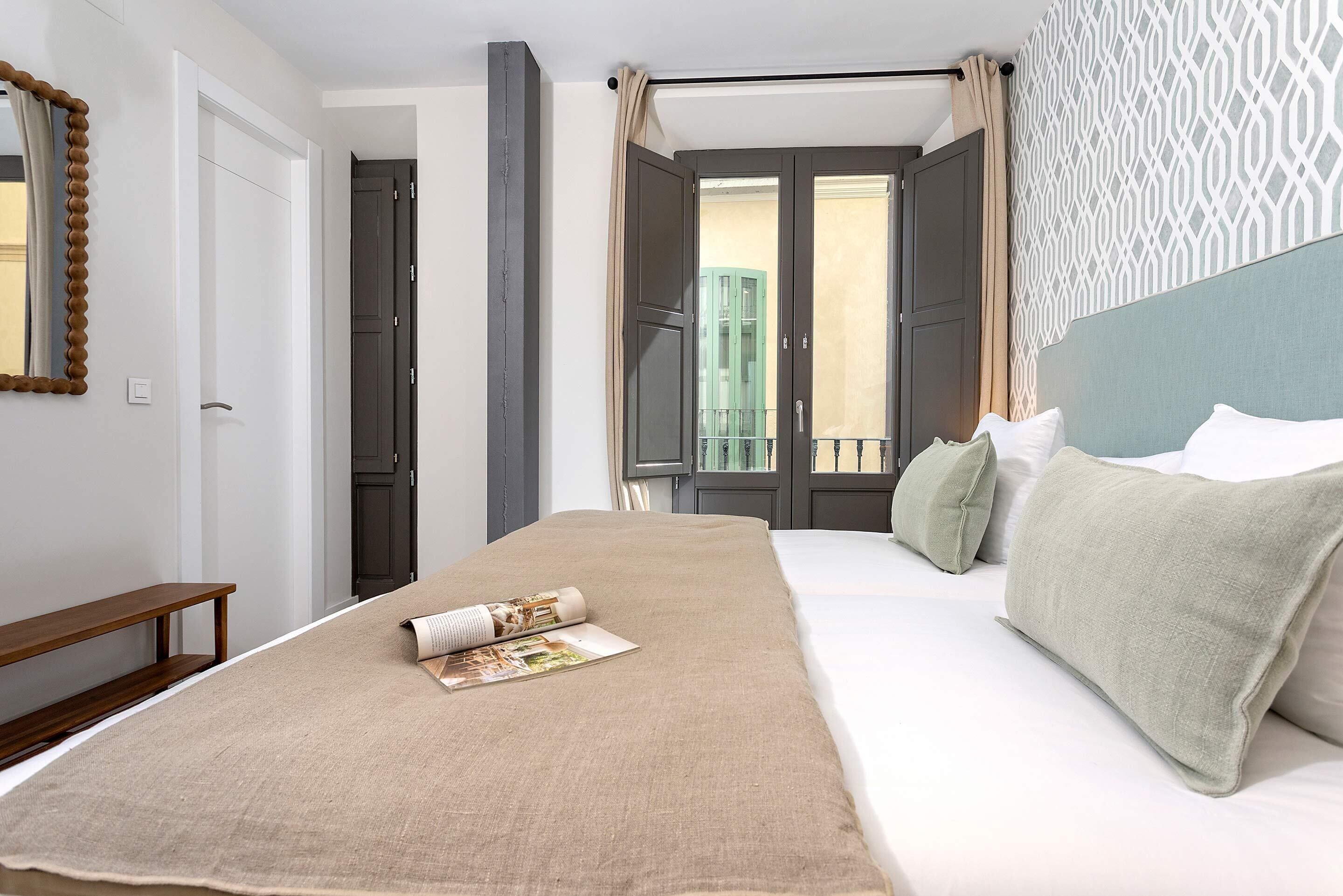 A hotel room with a large bed, a wooden bench, a mirror, and a view of the outside balcony.