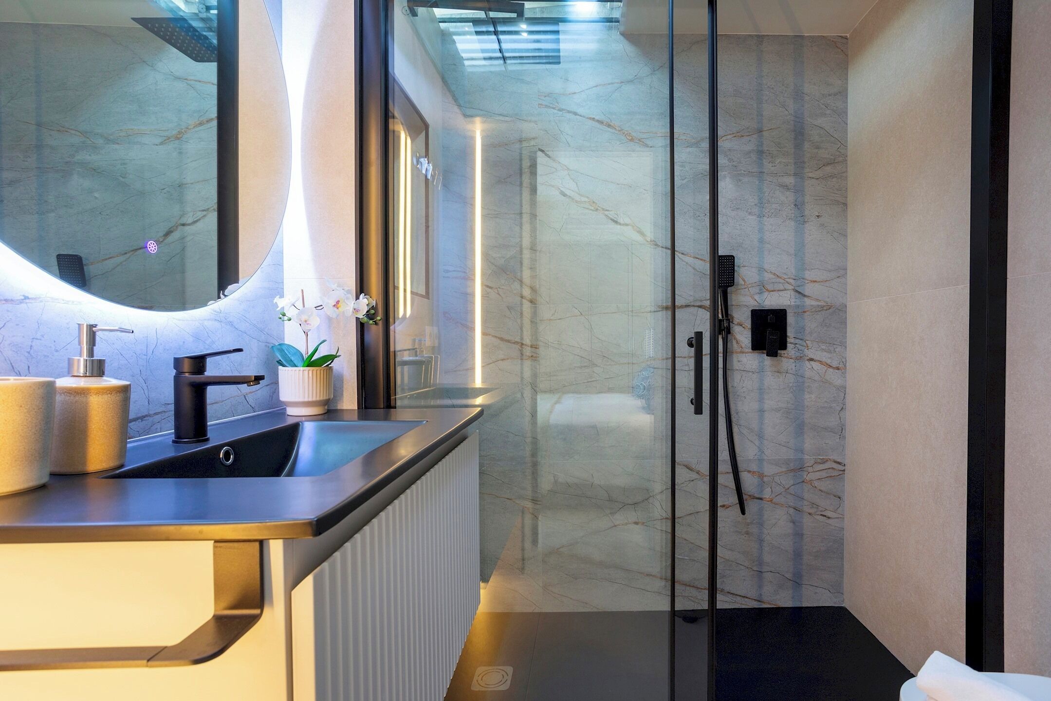Une salle de bain moderne dotée d’une douche à parois de verre, d’un mur recouvert de carreaux de marbre et d’un plan de toilette avec un lavabo et des objets décoratifs.
