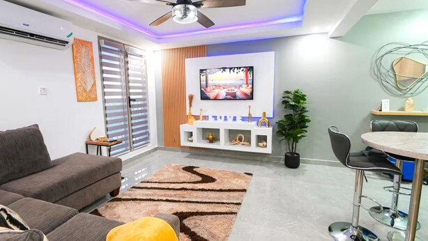 A modern living room with a sofa, a dining table with chairs, a ceiling fan, and a TV mounted on the wall. - Premium Suite at The Vineyards (Kingston)
