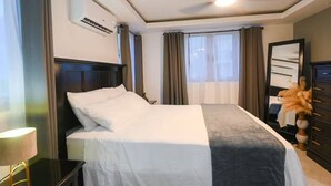 A hotel room with a bed, a ceiling fan, an air conditioning unit, a dresser, and a window with curtains. - Premium Suite at The Vineyards (Kingston)