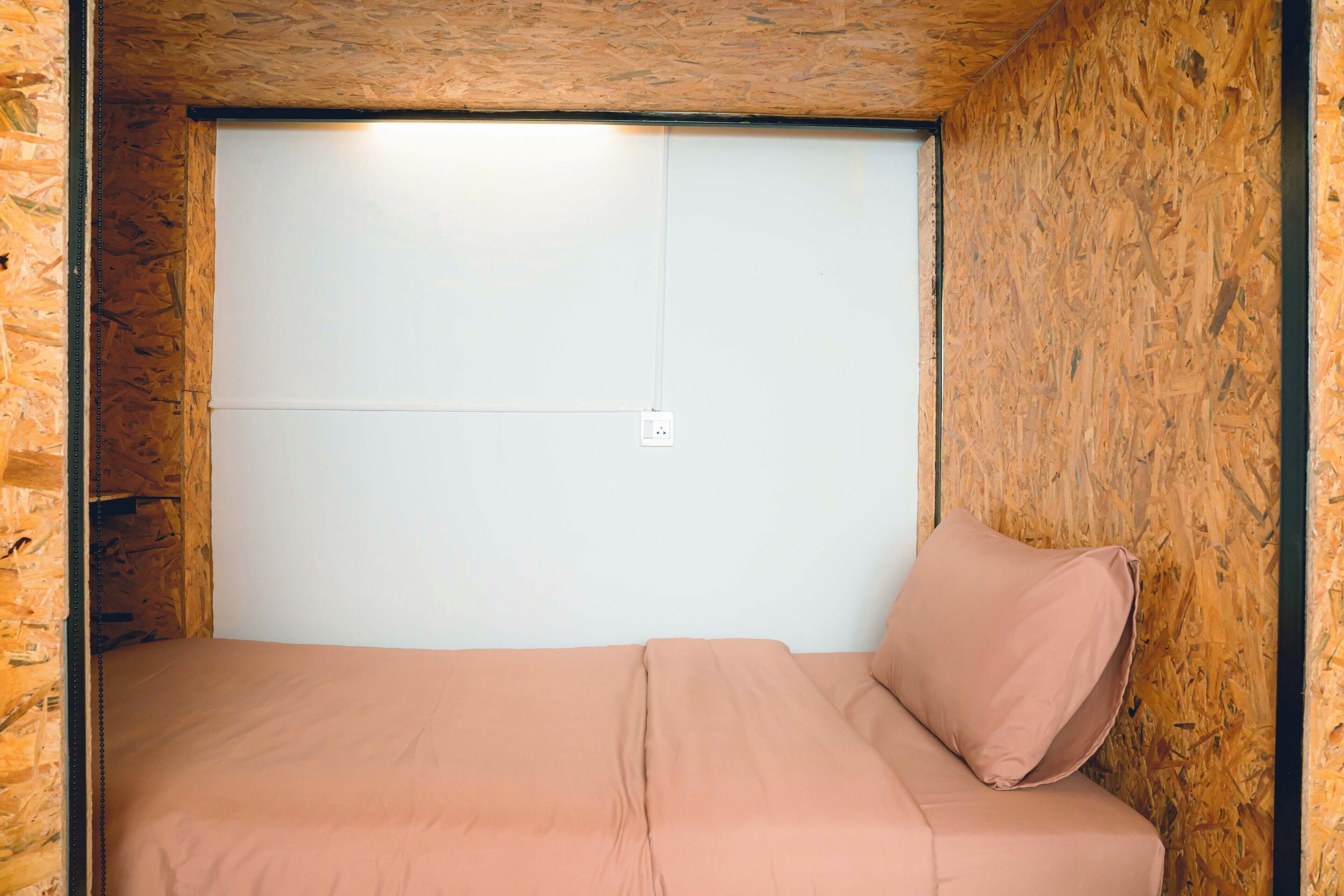 A small, single-bed room with a wooden ceiling and walls, a plain white door, and a single pink pillow.