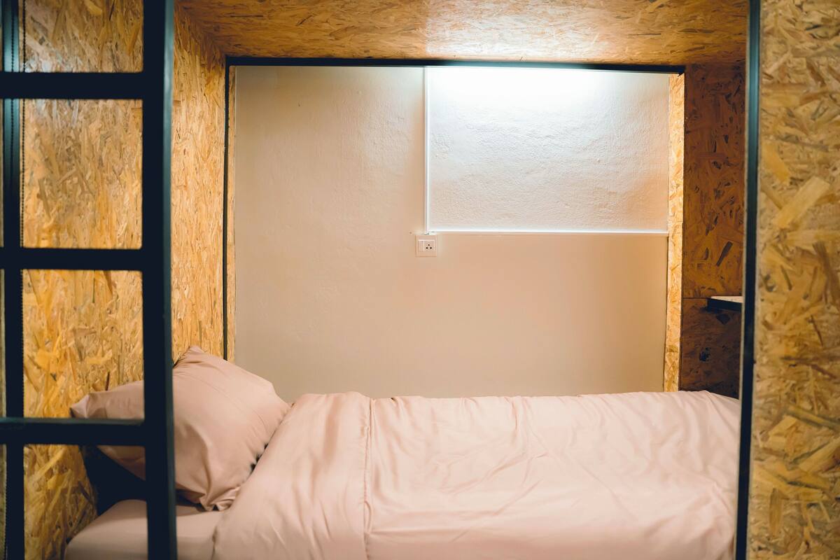 A bunk bed with a window above it, wooden walls, and a power outlet on the wall.