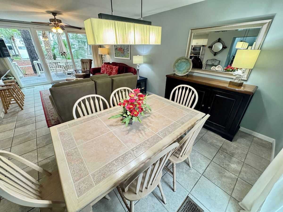 A dining area with a tiled table, chairs, and a vase of flowers. There is a sofa and a dining table in the background.