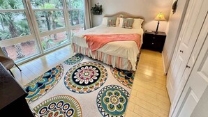 Un dormitorio con una cama grande, un ventilador de techo, un ventanal con vistas a la vegetación y una alfombra colorida.