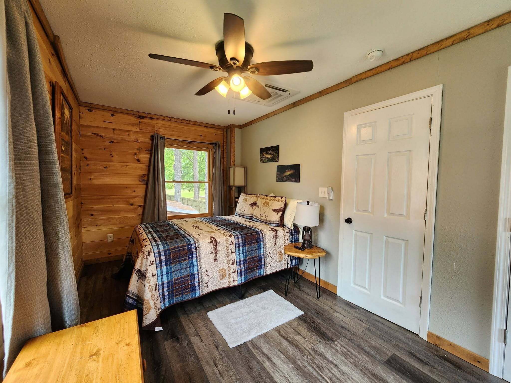 A room with a bed, a ceiling fan, a window, a door, and a small table with a lamp.