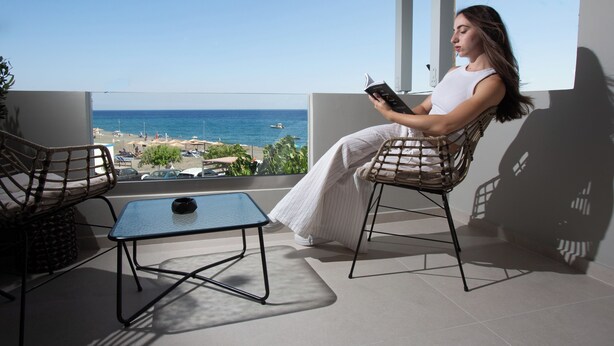 Un balcon avec vue sur la mer, deux fauteuils en osier, une petite table et une personne qui lit un livre.