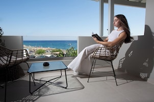 Un balcón con vista al mar, dos sillones de mimbre, una mesita y una persona leyendo un libro.