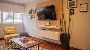 A modern living room with a large flat-screen TV mounted on a wooden wall, a comfortable yellow armchair, a coffee table with books, a purple ottoman, and a tall black planter with long green plants. - Sophisticated 1BR Stay in Prime Miraflores (Lima)