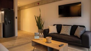 A modern living room with a black sofa, a wooden coffee table, a large potted plant, a flat-screen TV mounted on the wall, and a refrigerator in the background. - w Scenic 1BR w Terrace in Barranco (Lima)