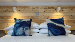 Quarto com cabeceira de madeira, roupa de cama branca e duas almofadas decorativas com desenhos abstratos.